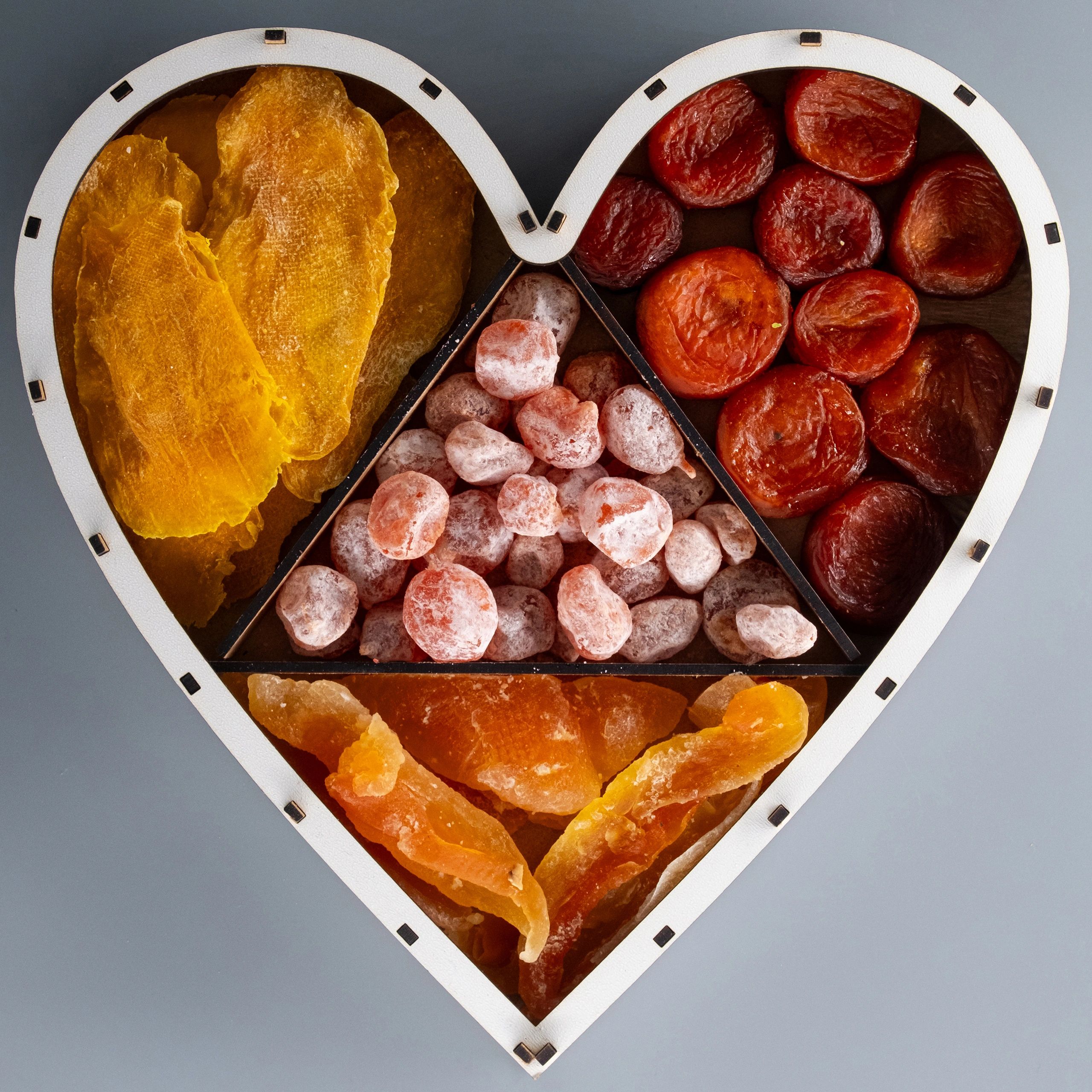 Various dried fruits in a heart-shaped wooden box. Variety of dried fruits are seen from above, including mango, kumquat, apricots, and cantaloupe.